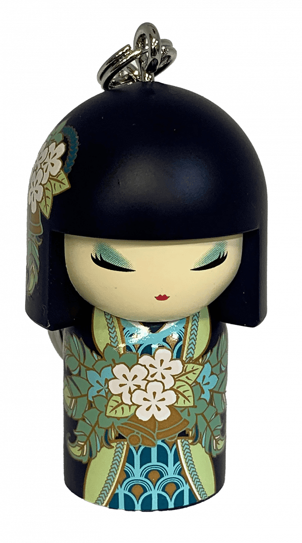 Un pequeño llavero con figura de una muñeca con cabello negro, ojos cerrados y patrones florales en su ropa, sosteniendo un ramo de flores blancas.